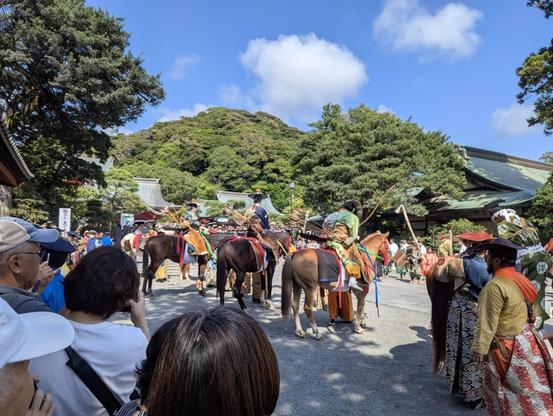 Une foule assiste à une procession traditionnelle japonaise : des cavaliers en costumes anciens richement colorés, avec arcs et carquois, attendent devant un sanctuaire entouré de collines boisées sous un ciel bleu ensoleillé.