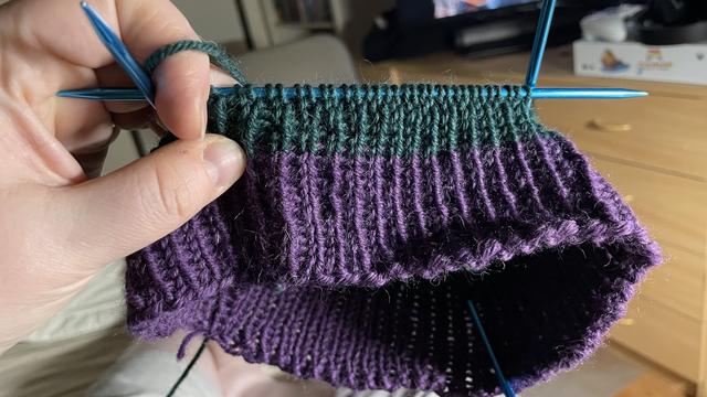 Neulotun raidallisen pipon alku. Puikot ovat siniset. Vasen käteni pitää työtä kameran edessä