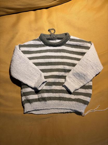 Knot sweater for a toddler. It is striped in khaki green and light blue, with light blue sleeves. 
The ends are still dangling on the sides.