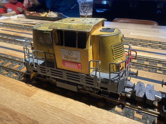 gelbe Modellbahnlokomotive auf Schienen, am Führhäuschen sind zwei Schilder befestigt, einmal das Logo Vytopna, einmal der Name Mariana, die Lok fährt nach links aus dem Bild, im Hintergrund ist ein Bierglas und ein längliches Essgefäß zu sehen
