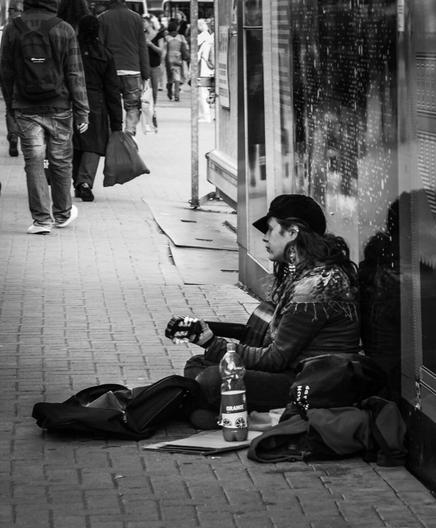 Na czarno-białej fotografii widać ulicznego muzyka siedzącego na chodniku, opartym o ścianę budynku. Osoba ma na sobie ciemną czapkę z daszkiem i kolczyki, trzyma gitarę, a obok niej stoją torby i butelka napoju gazowanego. Przed muzykiem leży otwarta torba, prawdopodobnie przeznaczona na datki. W tle widać przechodniów idących chodnikiem – większość z nich jest odwrócona plecami. Zdjęcie ma dokumentalny charakter, uchwycone zostało codzienne życie miejskiej ulicy, z nutą melancholii i samotności artysty grającego wśród obojętnych przechodniów.
