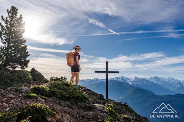 Woman with an orange backpack standing next to a summit cross. Beautiful mountains in the background. Tree on the left side.