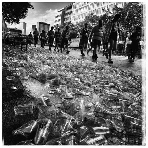 Street scattered with hundreds of discarded plastic cups, many labeled "MADE FROM RECYCLING MATERIAL," with a group of marathon runners or participants in a race walking and running in the background. Urban buildings and trees line the street, and the scene has a lively, chaotic atmosphere under daylight, rendered in black and white for dramatic effect.