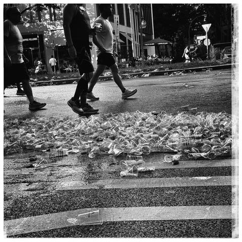 A large pile of discarded plastic cups scattered on the asphalt of a city street. Several people, likely runners or participants in an athletic event, are walking past the cups. The scene has an urban backdrop with buildings and pedestrians in the distance. The image is in black and white, creating a dramatic and gritty atmosphere. The sunlight creates shadows and highlights, enhancing the contrast in the photo.