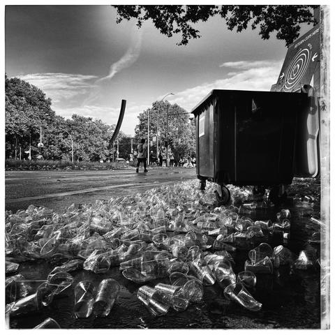 A messy urban street scene at the Berlin Marathon with hundreds of plastic cups scattered all over the wet pavement near a large wheeled trash bin. In the distance, a few people are walking away, possibly after an event or race. Surrounding the street are leafy trees and some urban elements like lamp posts and a billboard. The image is black and white, with high contrast between the shiny plastic cups, the dark bin, and the bright sky above.