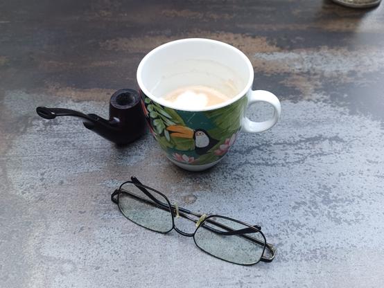 Eine bunt bemalte Kaffeetasse mit tropischem Motiv und einem Tukan steht auf einem grauen Tisch. Daneben liegen eine schwarze Brille und eine Pfeife.