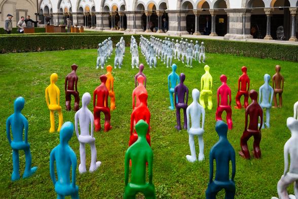 Im Vordergrund stehen bunte, menschliche, Figuren in Reih und Glied aufgestellten monochromen menschlichen Figuren, auf einer Wiese gegenüber.
Im Hintergrund sind die Säulen des Kreuzgangs der Abtei zu erkennen