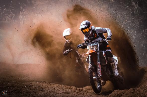 Landscape format photo. Two motocross riders battling it out in a race. The motorcycle on the right, slightly ahead, kicks up a lot of dirt, and the second rider is almost swallowed up in the cloud of dust.