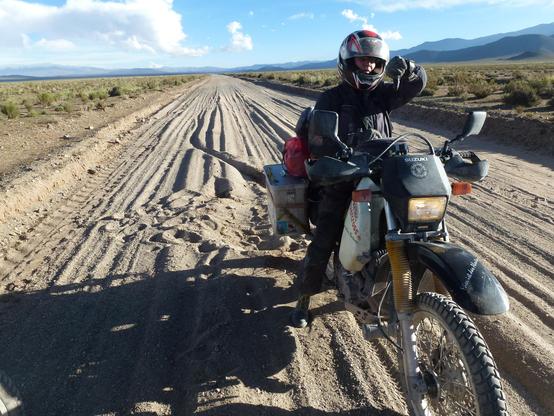 eine Verspurte Sandpiste führt in den Horizont. im Vordergrund eine stehende Motorradfahrerin die mit dem Daumen nach unten zeigt. kurz hinter ihr ihre schlingernde Fahrspur und ein 'Einschlagkrater'