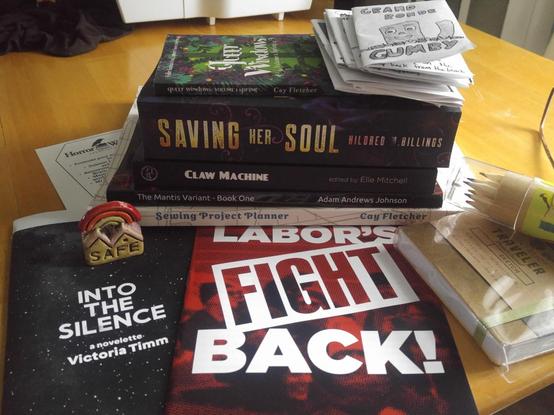 A stack of books, pamphlets, and zines. "Into the Silence" by Victoria Timm, "Labor's Fight Back!" from the Communist Party USA Labor Commission, "Sewing Project Planner" by Cay Fletcher, "The Mantis Variant - Book One" by Adam Andrews Johnson, "Claw Machine" edited by Elle Mitchell, "Saving her Soul" by Hildred M. Billings, "Queer Windows: Volume 1 Spring" by Cay Fletcher, and a stack of zines topped with "Grande Ronde Gumby." There is also a sketch book in shrink wrap with a tube of colored pencils, a card from the Horror Writers Association, and a tiny ceramic "Safe House" sculpture with a red and orange rainbow.