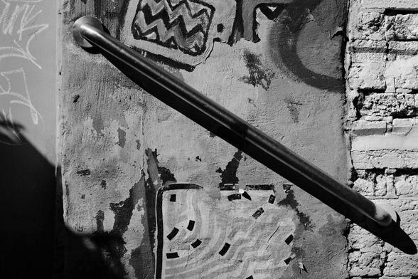 Black and white photo of a diagonal handrail attached to a wall in an alleyway. The wall consists of various surfaces, and has graffiti on it.