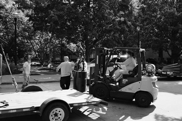 Black and white photo showing several people in the process of moving a new sculpture into Krutch Park in downtown Knoxville. It appears to be an abstract piece, and it is being moved from a trailer using a forklift.