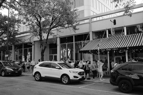 Black and white photo showing a long line of people along a sidewalk, waiting for their turn to get into a French crêpe restaurant downtown. It's not a new place, but is apparently very popular.