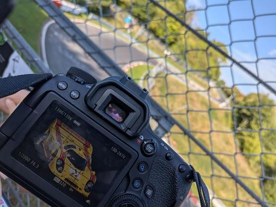 Im Hintergrund der Abschnitt WEHRSEIFEN auf der Rennstrecke. Kurvig und von der Sonne angestrahlt.
Vor einem Fotoloch im Zaun siehst meine Kamera mit einer Vorschau auf dem Display.