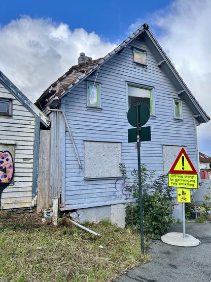 Two wooden houses next to Stavanger’s Nytorget square, both abandoned and in a poor state, windows sealed with wooden planks, some wild grown nature around, trash and a traffic sign. In the background blue skies and some clouds, it’s a sunny day