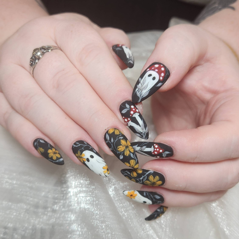 Long skinny almond black matte nails. The ring finger has a cute little ghostie, the pointer fingers have two amanita mushrooms, and the thumbs have a large amanita mushroom ghost with two black eyes. All of the nails are decorated with grey leaves and faded orange flowers.