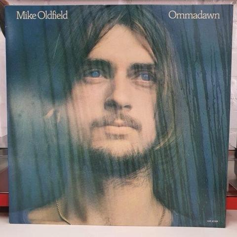 Album cover for "Ommadawn" by Mike Oldfield, featuring a close-up portrait of a young man with long hair, a beard and a thoughtful expression, set against a blue and green background.
