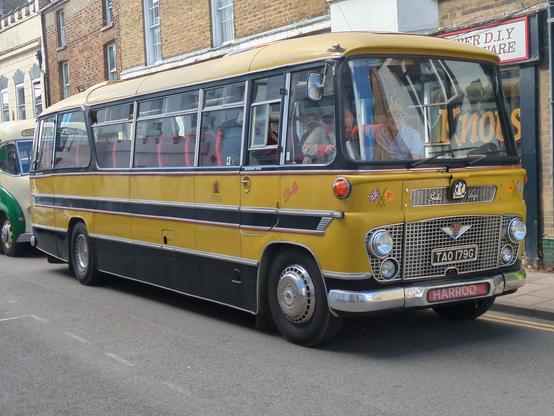 A mostly yellow 1960s Bedford coach, queuing in Downham Market with 18th century shop buildings just visible behind.
The coach has a lot of chrome, surrounding the windows and also the black side stripe and black lower quarter, as well as a large chrome front grille holding four lamps.