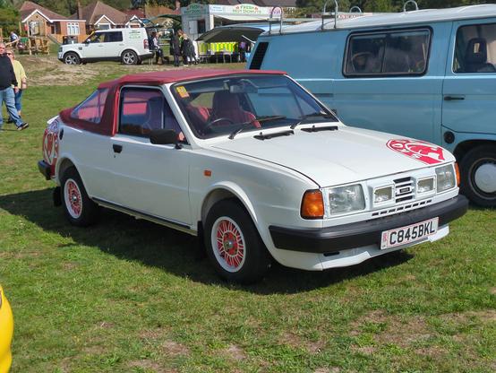 Where to begin... a white Skoda 130, complete with red, perhaps 15 inch diameter, vinyl Skoda badges on the front left edge of the bonnet and the back of the right rear wing. But there's so much more: white alloy wheels with red backgrounds between the spokes, a plastic/glassfibre add-on panel in place of the front grille (it's rear-engined) which contains two rectangular spotlights, and a red - possibly leather - hood over the convertible bodywork. The hood is a little ungainly, looking like a pram cover with soft flexible rear side windows.
It's parked in a classic car show field, next to a light blue VW van and with a burger stall in the background. It is absurdly magnificent.