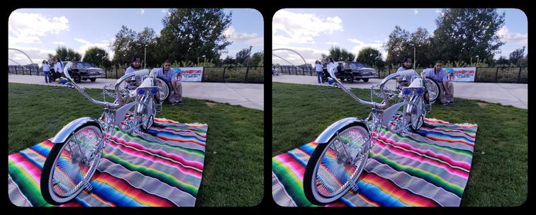 A chrome and grey lowrider-style bicycle standing on a colorful Mexican blanket on a lawn in a park. It has handlebars slanting forwards plus a steering wheel made of a chain. White banana style seat. Behind it are two young men in lawn chairs.
