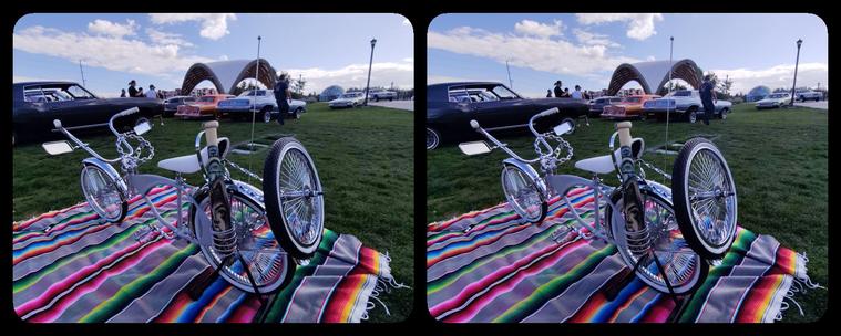 A view from the rear of the lowrider bicycle. There is a spare tire on the back and on the left side of the bike near the back tire, there is a holder containing what looks like a stubby baseball bat, decorated tattoo style.