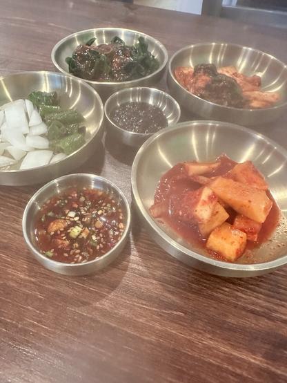 Multiple metal bowls with Korean food. There’s kimchi, turnip kimchi, seaweed, and onions with peppers. Plus bean paste and a chili sauce