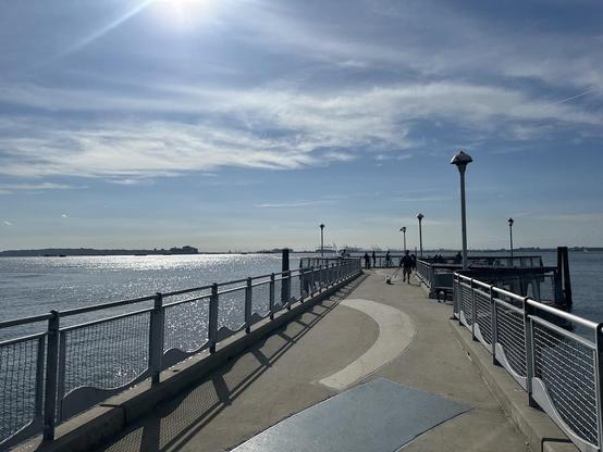 Photo looking along the length of Valentino Pier at Red Hook. It's a bit cloudy but still sunny. There is a cruise ship exiting New York Bay a mile or more away toward the left side of the photo, and another one docked at the Bayonne pierto its right.