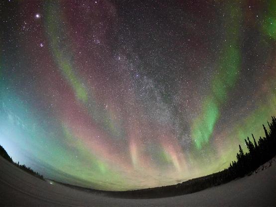 A photograph taken with a fish eye lens showing the Milky Way and the aurora borealis in the night sky above a frozen lake with a line of trees in the distant foreground.