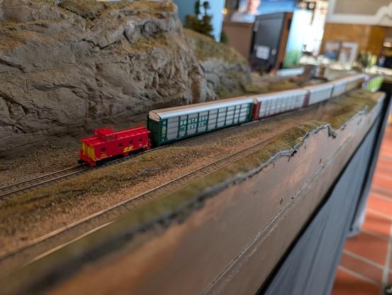 An N-scale autorack train goes down a long straight. There is a red Santa Fe caboose painted in the "Kodachrome" scheme