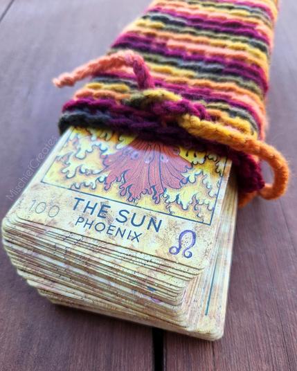 A crochet tarot card bag made from autumn-colored striped yarn. It's open, with a deck of cards sticking out. The card on top is The Sun.