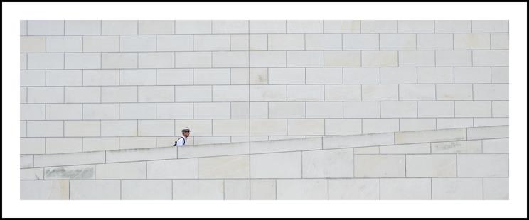 Das Bild zeigt einen Mann mit weißem Hemd und hellem Hut, der einen Rucksack trägt und eine leicht ansteigende, helle Steinmauer entlanggeht. Hinter ihm ist eine weitere hohe Mauer oder Fassade aus den gleichen Steinen. Der Mann ist nur vom Kopf bis etwas unterhalb der Schultern sichtbar. Der Rest ist hinter der vorderen Mauer versteckt, die von links nach rechts ansteigt. Die Mauern sind aus rechteckigen Steinen in hellen Creme- und Weißtönen gefertigt und dominieren das Bild. Der Mann ist im Vergleich zur Mauer klein und wirkt fast verloren in der Weite der Architektur. Der Fokus liegt auf der Einfachheit der Komposition und dem Kontrast zwischen der menschlichen Figur und der massiven Architektur.

The picture shows a man wearing a white shirt and a light-colored hat, carrying a backpack and walking along a slightly ascending, light-colored stone wall. Behind him is another high wall or facade made of the same stones. The man is only visible from the head to just below the shoulders. The rest is hidden behind the front wall, which rises from left to right. The walls are made of rectangular stones in light cream and white tones and dominate the image. The man is small in comparison to the wall and seems almost lost in the vastness of the architecture. The focus is on the simplicity of the composition and the contrast between the human figure and the massive architecture.