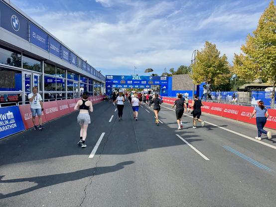 Eine geschäftige Szene an einer Marathon-Ziellinie. Läufer unterschiedlichen Alters und Fitnessniveaus überqueren den Zielbereich, gesäumt mit Werbebannern von Sponsoren. Zuschauer sind anwesend, und ein Bildschirm zeigt das Branding der Veranstaltung an. Die Umgebung ist sonnig mit Bäumen in