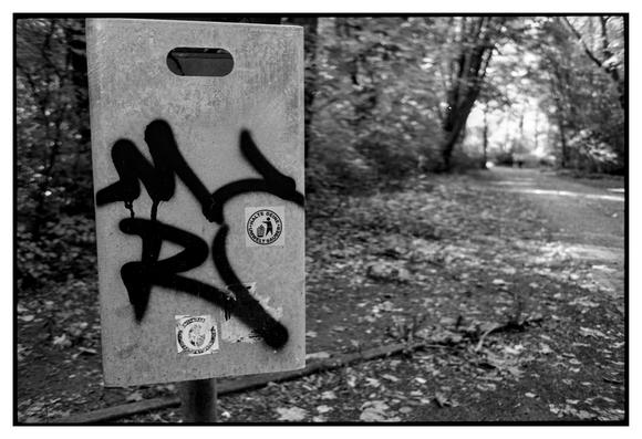 Black and white photograph in landscape orientation. A scene from a park with a dustbin in the foreground. The trees and paths in the background are out of focus. On the dustbin one can see some graffiti and a antifa sticker: A person throwing a swastika into a dustbin saying "Halte Deine Umwelt sauber".