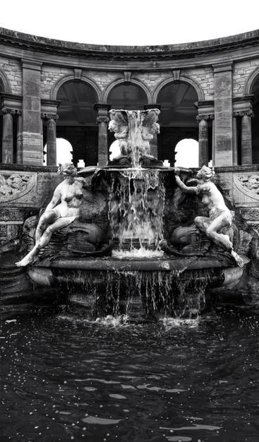 A dramatic, black and white, eye-level shot captures a grand classical stone fountain and colonnade. In the foreground, a dark pool of water, flecked with foam, reflects the scene. The ornate fountain features a central column from which water cascades forcefully, creating a dramatic, blurred effect. Two large, classical nude figures, carved from stone, recline on either side of the fountain's base. Behind the fountain, a semi-circular colonnade with multiple arches and pillars frames the upper part of the image, showcasing intricate architectural details and leading the eye upwards. The overall mood is timeless and imposing, enhanced by the monochrome palette.