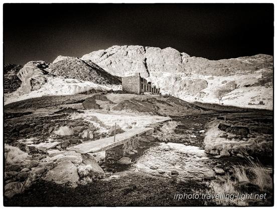 A toned black and white infrared photo of a disused slate mining area, with a clapper bridge over a stream in the foreground and a roofless terrace of living quarters in the background.