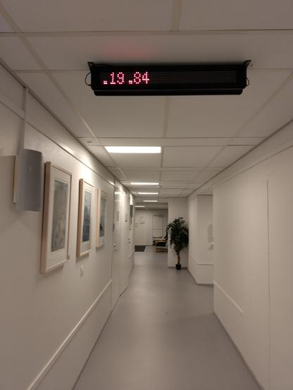 Käytävä, jonka katossa on  digitaalinen kellotaulu, jossa lukee punaisella .19 .84. 

Käytävä on vaalean sävyinen, seinillä sisustustauluja, joku iso tekokasvi taustalla.