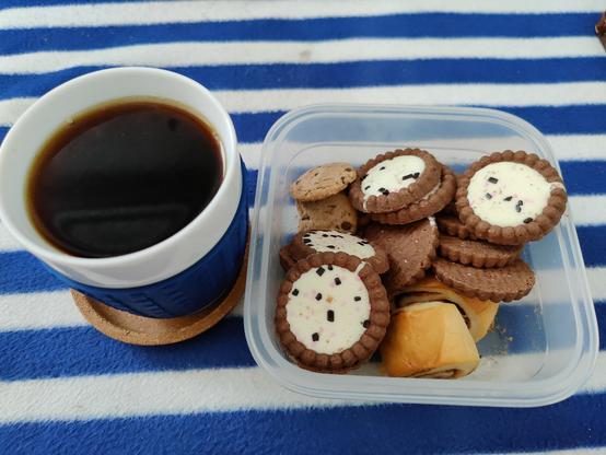 Täysinäinen valkoinen kahvimuki vieressä jämäboxi (Doris keksejä, muutama suklaapilkkukaurakeksi, muutama kuivakka pågenspulla)