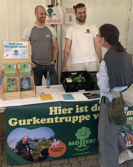 Das Bild zeigt einen Messestand von **Möllers Morgen** auf einer Veranstaltung. Zwei lächelnde Männer stehen hinter einem Tisch, auf dem Informationsmaterial, eine Kiste mit Gemüse und ein Schild mit der Aufschrift **"Teil von Morgen sein?"** zu sehen sind. Im Vordergrund steht auf einem grünen Banner:
**"Hier ist die Gurkentruppe von Möllers Morgen!"**
Die Szene wirkt einladend und informativ, als würden die beiden Besucher:innen über die Solidarische Landwirtschaft und die Arbeit von Möllers Morgen aufklären. Der Stand lädt zum Mitmachen und Informieren ein! 🥒🌱