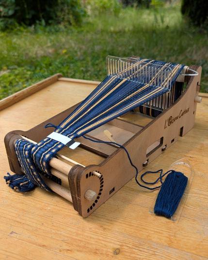 Photo d'un petit métier à tisser en bois posé sur une table dans un jardin. Sur le métier à tisser, un galon bleu avec des touches ocres est en cours de tissage au peigne d'encroix.
