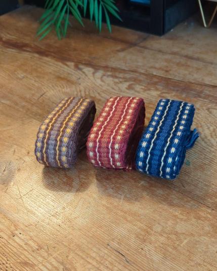 Photo de galons tissé roulés et posés sur une table en bois. 3 coloris : brun, rouge et bleu avec un motif de lignes et pointillés dans des fils ocre.