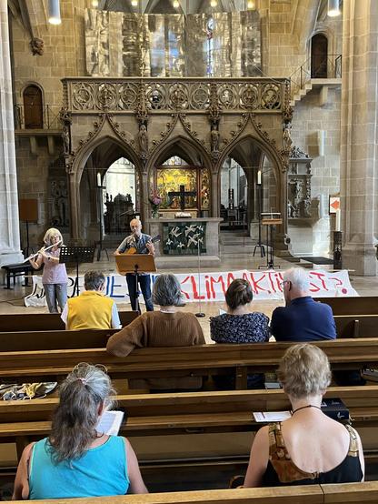Wenn schneiden einer Kirche in Tübingen. Vorne stehen zwei Musiker:innen. Hinter ihnen ist ein Banner mit der Aufschrift: Wäre Jesus Klimaaktivist?