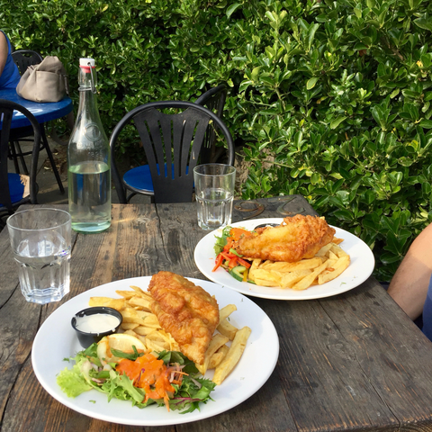 holztisch in der aussengastronomie … ein leerer stuhl … auf dem tisch zwei teller fish and chips, zwei gläser, eine wasserflasche …