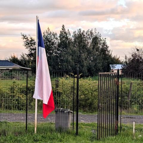 Una bandera chilena cuelga casi inmóvil de un mástil blanco, mostrando sus franjas verticales azul, blanca y roja. En primer plano, césped verde. Detrás del mástil, una cerca de malla metálica oscura se extiende a lo largo de la imagen, con una puerta de metal abierta a la derecha. Junto al mástil, hay un cubo de basura gris oscuro. Al fondo, se ve una densa línea de árboles verdes oscuros y, más allá, tejados de casas dispersas. El cielo está nublado con suaves tonos rosados y anaranjados de atardecer.