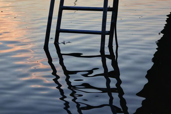 A high-angle, close-up shot captures the intriguing reflection of a ladder in rippling water during what appears to be dawn. The top half of a dark metal ladder, with three visible rungs, extends out of frame at the top. Its lower half and shadow are submerged, creating a distorted, elongated reflection in the gently undulating surface of the water. The water itself is a mix of deep blues and purples, with subtle patches of soft orange and pink reflecting from the sky on the left. The overall impression is serene and artistic, focusing on the interplay of light, shadow, and movement.