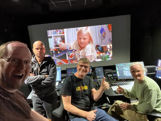Four people in a dark mixing theatre. From the left: happy man with glasses, close to the camera. Behind on the left: man in a hoodie, standing with arms crossed. Smiling man and woman, both with glasses, sitting in front of the mixing desk on separate chairs, both giving thumbs up. On the screen in the background: a girl playing with LEGO.