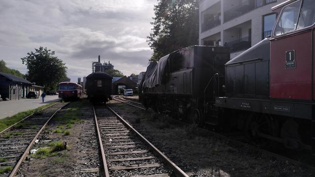 Bahnhof Kappeln Hafen, tagsüber, bedeckter Himmel. Drei Gleise mit leichtem Oberbau, entlang der Gleise fotografiert. Links ein Schienenbus, mittig Reisezugwagen, rechts am Bildrand eine kleine zweigekuppelte Henschel Stangendiesellok und davor eine große Tenderlokomotive. Achsfolge 1C2, großer Bunker über dem Drehgestell, und entgegen deutscher Praxis sind auch Rahmen und Fahrwerk schwarz. Führerstand und Kessel sind unter einer Plane.
Zwischen Reisezugwagen und Dampflok ist der einständige freie Lokschuppen zu sehen, vorderes und hinteres Tor offen.
