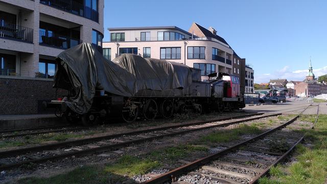 Dampflok und Diesellok von schräg vorne. Fahrwerk ist besser sichtbar, an der Walschaert Steuerung fehlen die Kuppelstangen, Treibstange, Gegenkurbel, und Schwingenstange. Direkt hinter Gleis und schmalem Zuweg stehen moderne und uninspieriert fancy Wohngebäude mit Ziegeltapete, große zurückgesetzte Balkone und bodentiefe Fensters.