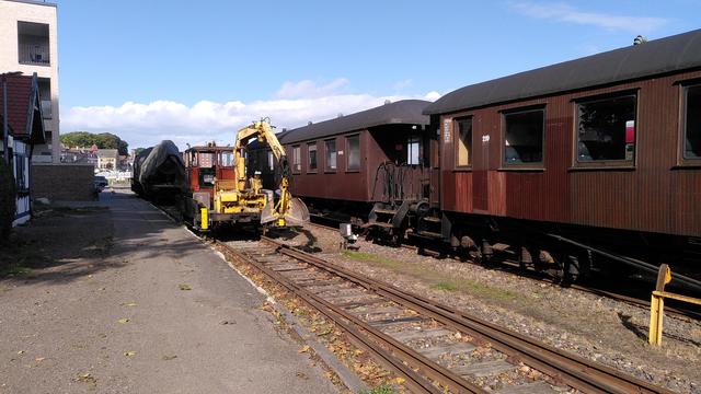 Blick von der Weiche zum Lokschuppen Richtung Dampflok. An der Spitze der Weiche steht jetzt ein doitsches Skl Klv53, ein kleines zweiachsiges Baufahrzeug mit orangener Kabine, Ladefläche, und gelbem Hydraulikkran, daran eine Baggerschaufel. Auf dem mittleren Gleis rechts daneben die klar lackierten Drehgestell-Personenwagen, die sind, wie die Dampflok, aus Skandinavien.