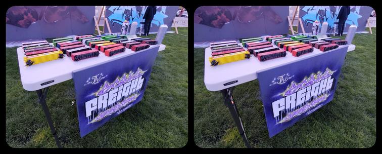 On a small table at an outdoor arts festival, a dozen or so model train freight cars and some cans of spray paint. The purple banner in front is lettered in graffiti style "The Scale Freight Connection."