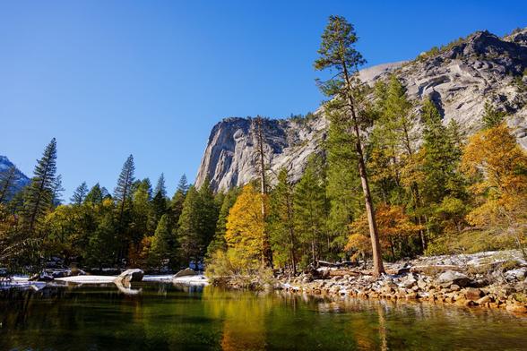 A tranquil river flows through a forest, with colorful autumn leaves and rocky cliffs under a clear blue sky.